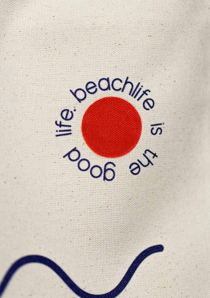 Close-up of a cotton tote bag with the beachlife is the good life message placed around a small orange circle.