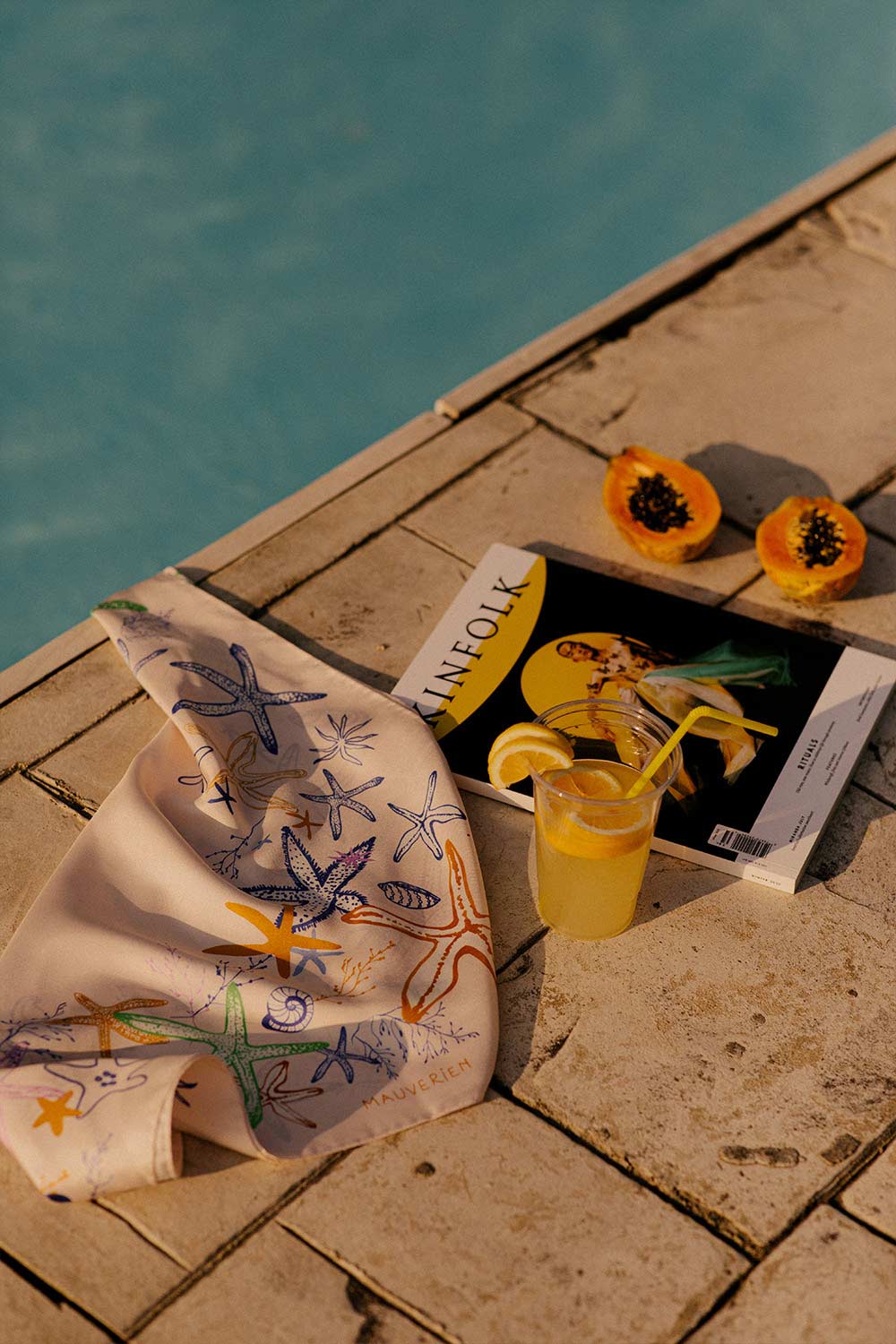 Beige silk scarf with starfish laid by the pool with a glass of juice and a magazine.